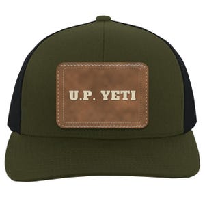 May include: An olive green baseball cap with black side panels and a rectangular brown leather patch centered on the front. The patch features the text U.P. YETI in a bold, light-colored font with visible stitching around the edges.