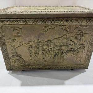 May include: A rectangular brass storage box featuring an embossed relief scene of a horse-drawn carriage, figures, and a building on the front panel. The lid and edges are decorated with a repeating geometric pattern in a metallic finish.