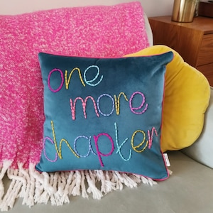 Op de afbeelding: Teal vierkant kussen met de geborduurde zin "one more chapter" in kleurrijke draad. Het kussen heeft een roze rand en ligt op een grijze bank naast een roze deken met witte franjes en een geel kussen.