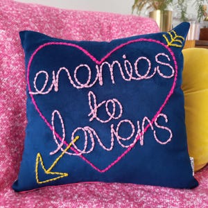 Op de afbeelding: Een marineblauw vierkant kussen met de geborduurde zin "enemies to lovers" in een roze hart. Een gele pijl en bladeren accentueren het ontwerp. Het kussen ligt op een roze oppervlak met patronen.