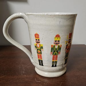 May include: A cream-colored ceramic mug with a speckled finish and a large handle. The exterior features a repeating graphic print of three nutcracker figures in red, green, and yellow uniforms, each wearing a tall hat and standing in a vertical row.