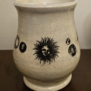 May include: A speckled, off-white ceramic mug with a flared top and a rounded base. The mug features black illustrations of celestial bodies, including a sun with a face. The mug is approximately 5 inches tall.