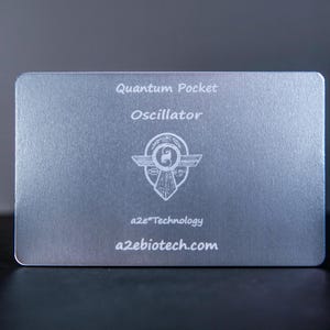 May include: A rectangular silver-toned metal card featuring the engraved text Quantum Pocket Oscillator, a central circular emblem with wings, and the text a2e Technology and a2ebiotech.com printed in white on the front surface.