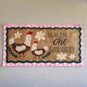 May include: A decorative sign featuring two cartoon chickens with red combs and white and brown bodies. The sign has the text "Islah June is... One Cutie Chick!!" surrounded by daisies and a pink and white striped border.