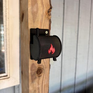 May include: A black, cylindrical wall-mounted holder featuring a red flame emblem on the front. The object is secured to a vertical wooden post with a single visible screw at the top, and it includes a small chimney-like protrusion on the top.
