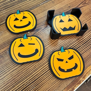 May include: A set of four orange pumpkin-shaped coasters with black facial features and teal stems. Three coasters are laid out individually, while one is stored inside a black plastic holder. Each coaster features a unique jack-o'-lantern expression.