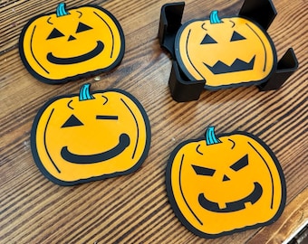 May include: A set of four orange pumpkin-shaped coasters with black facial features and teal stems. Three coasters are laid out individually, while one is stored inside a black plastic holder. Each coaster features a unique jack-o'-lantern expression.