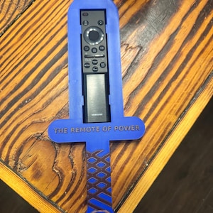 May include: A blue plastic sword-shaped remote control holder featuring a brown cross-hatched handle design and the text THE REMOTE OF POWER embossed on the crossguard. A black Samsung remote control is inserted into the central slot of the holder.
