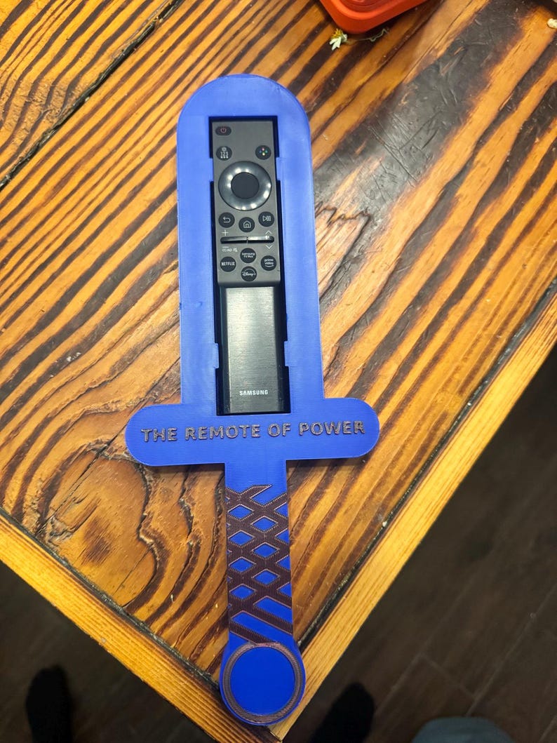 May include: A blue plastic sword-shaped remote control holder featuring a brown cross-hatched handle design and the text THE REMOTE OF POWER embossed on the crossguard. A black Samsung remote control is inserted into the central slot of the holder.