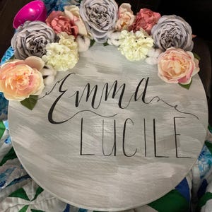 May include: A round, gray wooden sign with the names "Emma Lucile" in black script. The sign is decorated with a floral arrangement of pink, peach, white, and gray flowers. A pink toy is visible in the upper left corner.