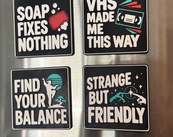 May include: Eight square black magnets with white text and colorful graphic icons. The text includes phrases like Raised on Cult Classics, Reality Needs a Reboot, Soap Fixes Nothing, VHS Made Me This Way, Find Your Balance, Strange But Friendly, Phone Home Later, and No Mercy for Mondays.