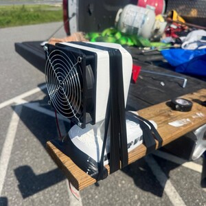 May include: A white 3D-printed housing unit featuring a black circular cooling fan with a silver wire grill. The device is secured to a wooden board with thick black rubber bands, with exposed red and black electrical wires extending from the base.