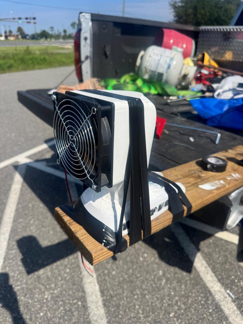 May include: A white 3D-printed housing unit featuring a black circular cooling fan with a silver wire grill. The device is secured to a wooden board with thick black rubber bands, with exposed red and black electrical wires extending from the base.