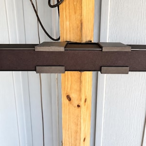 May include: A horizontal dark brown rectangular bar with four metallic clips attached to its front and top edges. The bar is mounted against a vertical light-colored wooden post, with a thin black electrical cord hanging down from the top area.