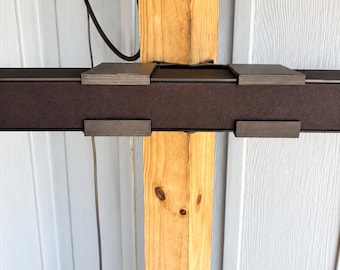 May include: A horizontal dark brown rectangular bar with four metallic clips attached to its front and top edges. The bar is mounted against a vertical light-colored wooden post, with a thin black electrical cord hanging down from the top area.