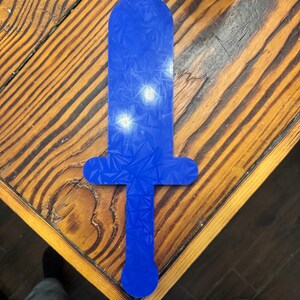 May include: A flat, blue, sword-shaped object featuring a geometric, crystalline holographic pattern across its entire surface. The item has a rounded tip, a crossguard, and a straight handle ending in a circular pommel, all in a single piece.