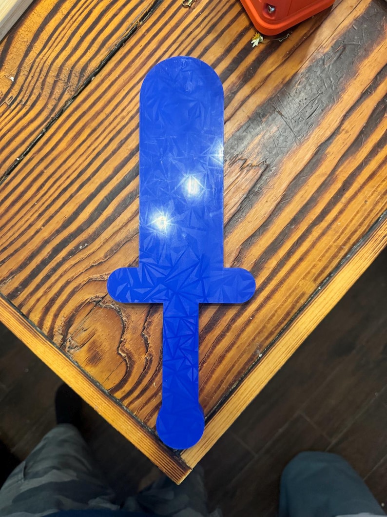 May include: A flat, blue, sword-shaped object featuring a geometric, crystalline holographic pattern across its entire surface. The item has a rounded tip, a crossguard, and a straight handle ending in a circular pommel, all in a single piece.