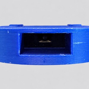 May include: A circular blue plastic component featuring a central rectangular recessed opening. Inside the dark rectangular cavity, a small metallic contact point is visible. The exterior surface of the blue plastic shows a textured finish.