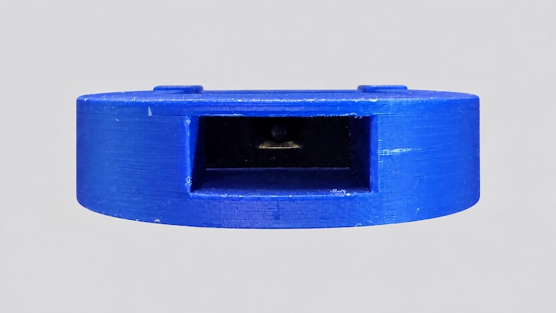 May include: A circular blue plastic component featuring a central rectangular recessed opening. Inside the dark rectangular cavity, a small metallic contact point is visible. The exterior surface of the blue plastic shows a textured finish.