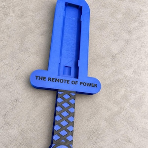 May include: A blue plastic sword-shaped remote control holder featuring a black cross-hatched grip and a circular pommel. The crossguard is printed with the black text THE REMOTE OF POWER, and the blade section has a recessed rectangular channel.