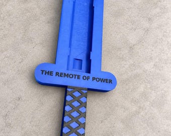 May include: A blue plastic sword-shaped remote control holder featuring a black cross-hatched grip and a circular pommel. The crossguard is printed with the black text THE REMOTE OF POWER, and the blade section has a recessed rectangular channel.