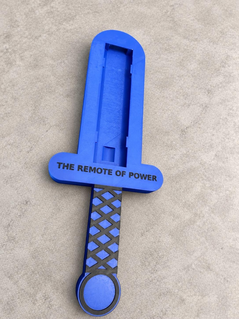 May include: A blue plastic sword-shaped remote control holder featuring a black cross-hatched grip and a circular pommel. The crossguard is printed with the black text THE REMOTE OF POWER, and the blade section has a recessed rectangular channel.