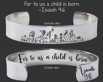 May include: A silver cuff bracelet with the text "For to us a child is born -Isaiah 9:6". The outside of the bracelet features a black line drawing of a nativity scene. The inside of the bracelet also has the text "For to us a child is born" and "Isaiah 9:6".