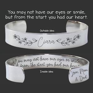 Könnte beinhalten: Silbernes Armreif-Armband mit einem floralen Design und dem Namen "Ciara" auf der Außenseite graviert. Die Innenseite des Armbands ist mit der Botschaft "You may not have our eyes or smile, but from the start you had our heart. Love, Mom & Dad." graviert.