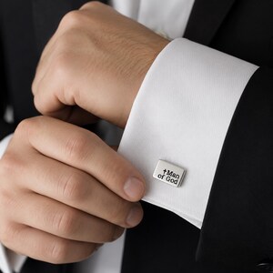 A close-up image of a person's hand adjusting a cufflink with the text "Man of God" engraved on it.