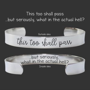 May include: A silver-toned metal cuff bracelet featuring two views. The exterior is engraved with the text 'this too shall pass' in a cursive script. The interior is engraved with the text '...but seriously, what in the actual hell?' in a sans-serif font.