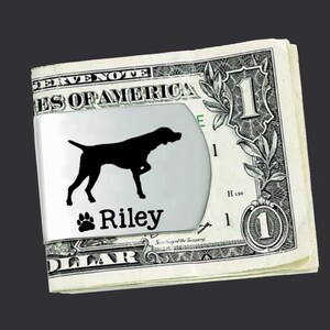 May include: A silver money clip with a black silhouette of a German Shorthaired Pointer dog and the name "Riley" engraved on it. The clip is holding a US dollar bill.