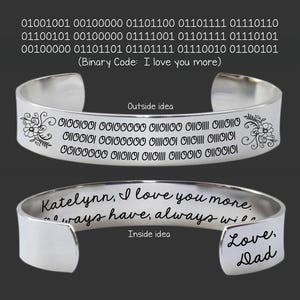 Op de afbeelding: Een zilveren manchetarmband met gegraveerde tekst en bloemmotieven. De buitenkant heeft binaire code en de binnenkant zegt "Katelynn, I love you more, always have, always will." Er staat ook "Love, Dad."
