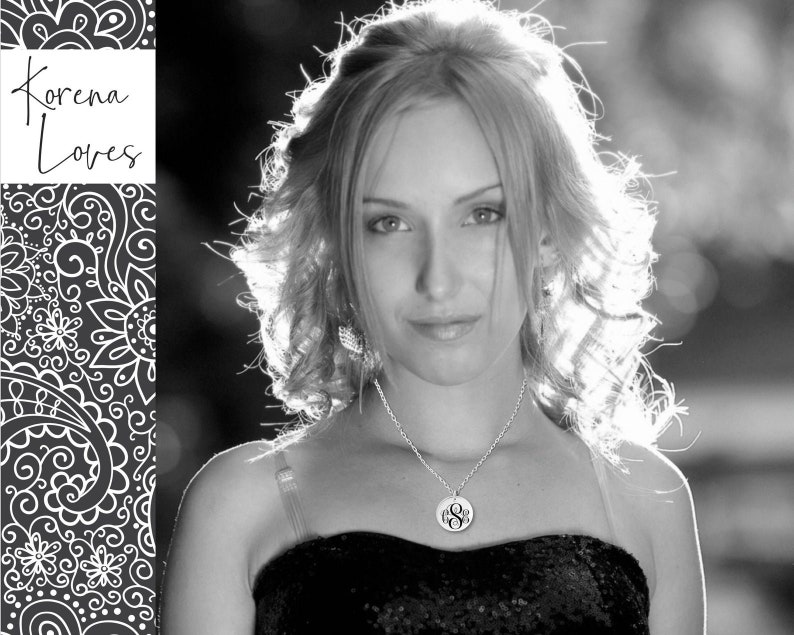 May include: A black and white portrait of a woman with long, wavy hair. She is wearing a black dress and a silver necklace with a monogram pendant.