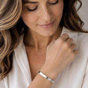 A woman with long, wavy brown hair wearing a white shirt and a silver bracelet is smiling and looking down at her hand.