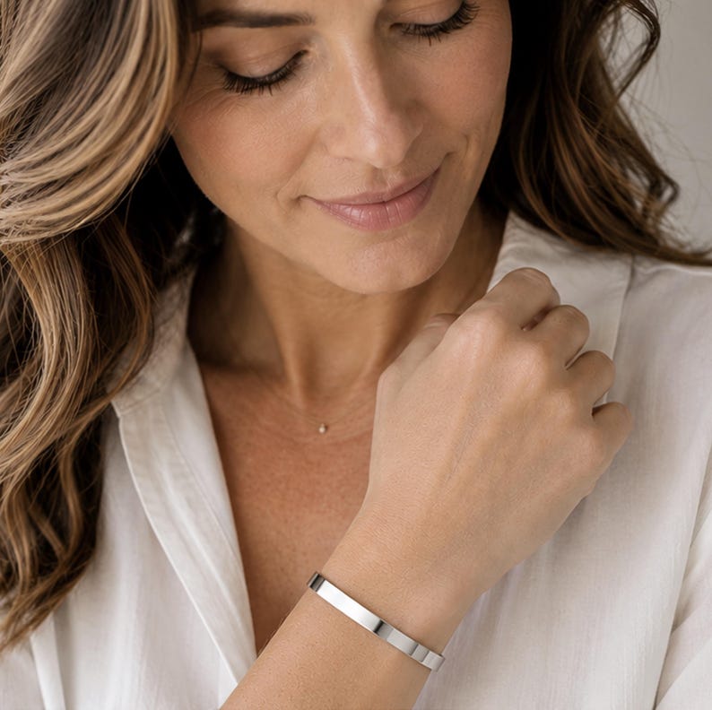 A woman with long, wavy brown hair wearing a white shirt and a silver bracelet is smiling and looking down at her hand.