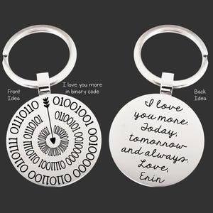 Op de afbeelding: Een zilveren sleutelhanger met een ronde hanger. De voorkant van de hanger heeft een ontwerp met een pijl die naar een cirkel van binaire code wijst. De achterkant van de hanger heeft de tekst "Ik hou meer van je. Vandaag, morgen en altijd. Liefs, Erin".