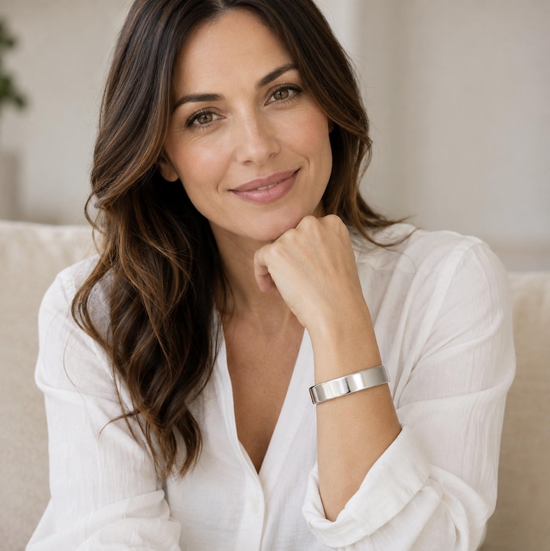 A woman with long brown hair wearing a white blouse and a silver bracelet is smiling at the camera.
