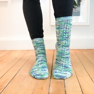 May include: A pair of colorful knit socks with a mix of blue, green, and purple hues. The socks are worn with black pants and are displayed on a wooden floor. The socks have a textured pattern.