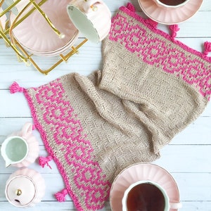Op de afbeelding: Een beige gebreide theekos met roze bies en kwastjes. De theekos is gedrapeerd over een theekopje en schotel met een roze bloemenpatroon. Twee extra theekopjes en schotels staan in een gouden metalen standaard.