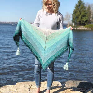 May include: A teal and green gradient shawl with a fringe on each end. The shawl is being held by a person wearing jeans and a grey sweater.
