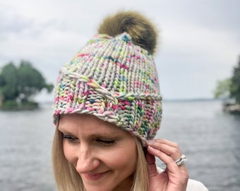 May include: A gray knit beanie with a colorful yarn pattern and a brown faux fur pom pom.