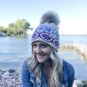 May include: A woman wearing a white, blue, and purple knitted hat with a fluffy pom pom. The hat has a geometric pattern. She is wearing a blue denim jacket and smiling.