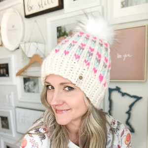 May include: A white knit beanie with a pink, blue, and gray chevron pattern and a white pom pom. The beanie has a small brown leather tag.