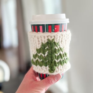 May include: A white knitted coffee cup cozy with a green knitted Christmas tree design. The cozy is on a red and green striped coffee cup.