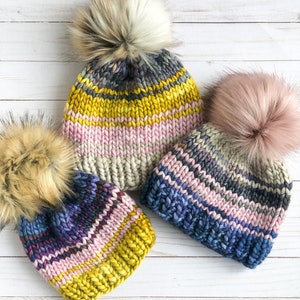 May include: Three hand-knitted beanies with faux fur pom-poms. The hats feature horizontal stripes in various colors, including yellow, pink, blue, and gray. The pom-poms are in shades of beige and pink, adding a touch of luxury to the winter accessories.
