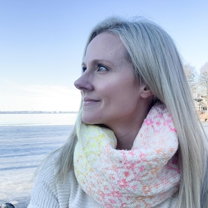 Op de afbeelding: Een vrouw met lang blond haar draagt een witte gebreide oneindige sjaal met een roze en gele ombre-ontwerp. Ze staat buiten met een waterpartij op de achtergrond.