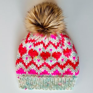 May include: A knitted hat with a pink, white, and blue heart pattern and a brown faux fur pom pom.