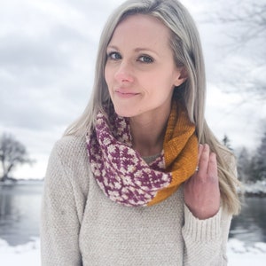 May include: A woman wearing a white sweater with a colorful infinity scarf. The scarf has a geometric pattern in shades of pink, white, and yellow.