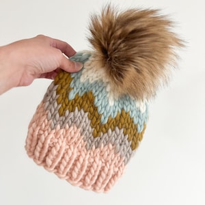 May include: A chunky knit hat with a large, fluffy brown pom-pom. The hat features a chevron pattern in shades of pink, gray, mustard yellow, light blue, and white. The hat is held by a hand against a white background.