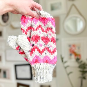 May include: A pair of pink, white, and gray knitted mittens with a chevron pattern. The mittens are held in a hand.
